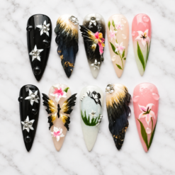 Handmade 3D butterfly and floral press on nails featuring black and gold butterfly wings, pink lilies, white flowers, rhinestones, and glossy gel details, displayed on white marble background.