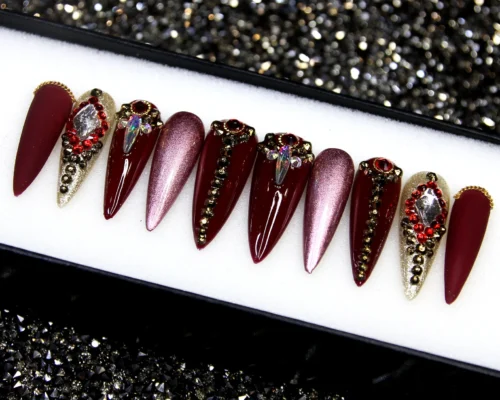 Oxblood and rose-gold chrome stiletto press-on nails in a tray, with red crystal clusters and bronze studs