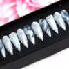 Grey marble stiletto press-on nails in a black tray beside pink flowers, with silver feathered ridges and pearls