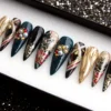 Angled view of black, teal and gold press-on nails, showing gold foil flowers, a red bloom and gem clusters