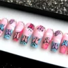 Pink coffin press-on nails with blue tips, black-outlined butterflies in blue, lilac and pink, and rhinestones, in a tray