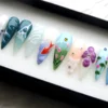 Teal, lilac and pale blue stiletto press-on nails in a tray, with a painted orange koi, pink tulips, a purple flower, waves