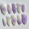 Lavender and white almond press-on nails flat on grey, with hand-painted purple flowers, a tiny bow, rhinestones and ombré