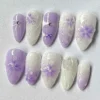 Mirrored flat-lay of white-to-lilac press-on nails, showing painted violet blossoms, glossy domes and a small purple bow