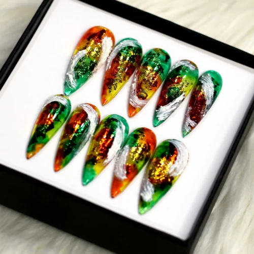 Green and orange marbled stiletto press-on nails in a black box on white fur, angled to show foil flecks and white swirls