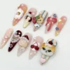 Long pink stiletto press-on nails flat on white: a ginger and white cat face, a swirl roll cake, a pink bow and pearl trims