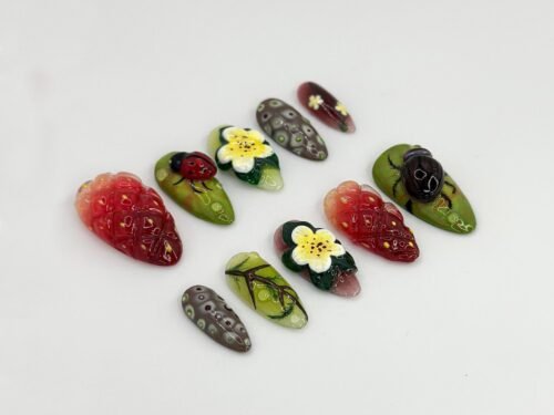 Green and red press-on nails on a white backdrop with raised strawberries, a ladybug, a dark beetle and a yellow flower