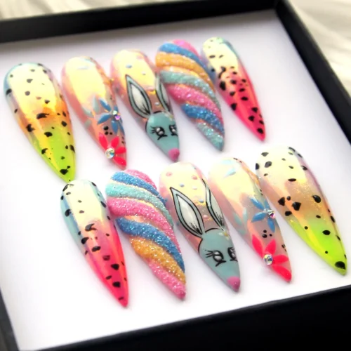 Bunny press-on nails seen at an angle in the box, with sugar-glitter rainbow stripes, blue flowers and pink-to-orange ombré