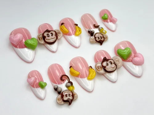 Pink almond press-on nails with white French tips, sculpted brown monkey faces, bananas and pink and green hearts, laid flat