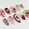 Almond press-on nails flat on white with painted strawberries, red and pink gingham, white polka dots and a cherry-topped bow
