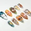 Long stiletto press-on nails on white with hand-painted cat faces, orange paw nails and a teal cat nail