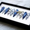 Navy chrome and grey press-on nails in a black box seen from the other side, with blue rhinestones and chain details