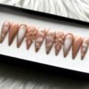 Caramel nude stiletto press-on nails in a tray on white fur, with milky marble swirls, gold foil edges and 3D flowers