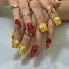 Press-on nails worn on a hand with gold rings: yellow sculpted corn-cob nails and red crawfish nails with black bead eyes