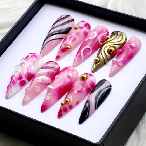 Angled box view of pink marble stiletto press-on nails with black striped accents, water-drop dots and a sculpted gold nail
