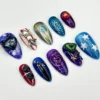 Purple, blue and red glitter almond press-on nails flat on white, with black 3D cats, a silver planet, spirals and stars