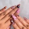 Press-on nails worn on a hand: cocoa brown and pink almond nails with white stars, polka dots, plaid and a 3D black charm