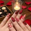Close-up of deep red and peach press-on nails with swirl patterns, held near rose petals and a candle