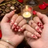 Sheer peach press-on nails with red bows, lattice and checkerboard worn on two hands, with a candle and chocolates