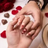 Peach press-on nails with dark red hearts and gingham worn on a hand, beside roses and heart chocolates