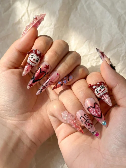 Clear pink stiletto press-on nails worn on two hands, with painted jester faces, hearts and tiny gems