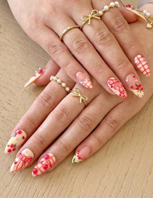 Peach almond press-on nails with pink gingham, painted bows and cherries worn on two hands with gold bow rings