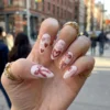 Milky white press-on nails with red hearts and a ribbon bow worn on one hand with gold rings, outdoors in the city