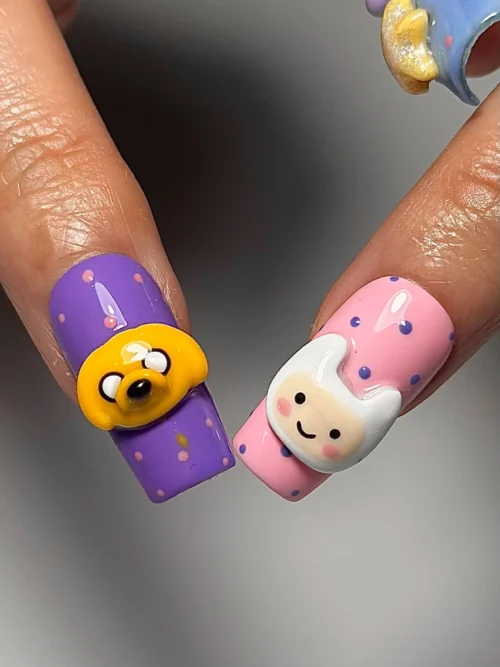 Close-up of two square press-on nails worn on a hand: a purple nail with a 3D yellow dog face and a pink 3D white-hooded face