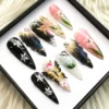 Angled view of black and pink press-on nails with feathered wing textures, a gold-and-black butterfly and painted lilies