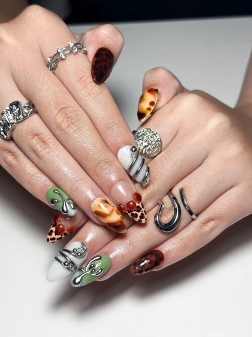 Amber and moss green press-on nails worn on two hands with silver rings, showing chrome swirls and leopard tips