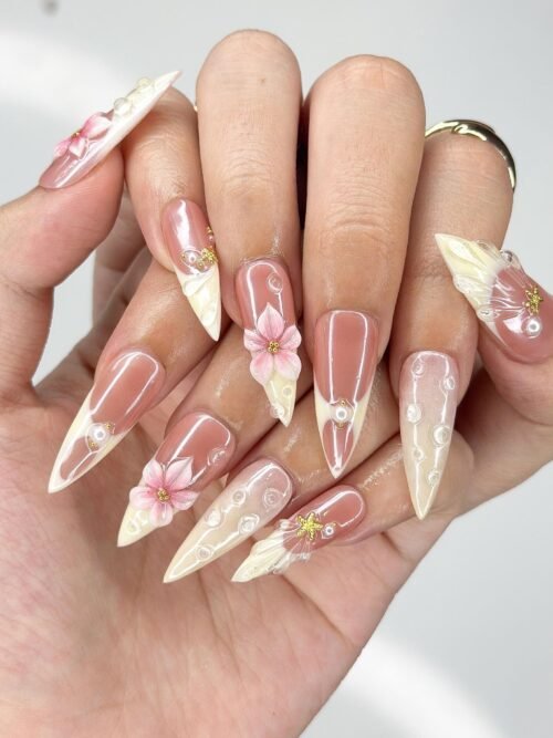 Peach stiletto press-on nails worn on a hand, with cream French tips, sculpted pink lilies and tiny pearls