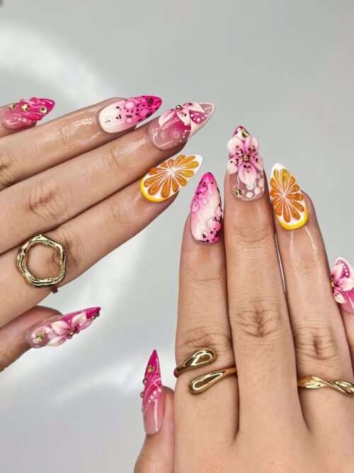 Fingers from another angle wearing pink press-on nails with orange slices and 3D pink lilies, with gold rings