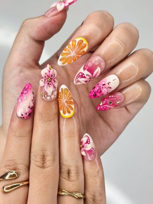 Pink almond press-on nails worn on a hand with sculpted orange slices, pink lilies and dark speckles
