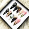 Black, blush and white stiletto press-on nails in a tray on white fur, with silver star flowers, painted lilies, a butterfly