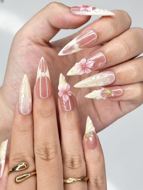 Worn on two hands with gold rings: nude pink stiletto press-on nails with cream tips, 3D pink flowers and tiny gold charms