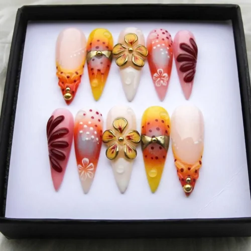 Orange-yellow ombre stiletto press-on nails in a black gift box, with gold flower charms, dots and wine-red petals