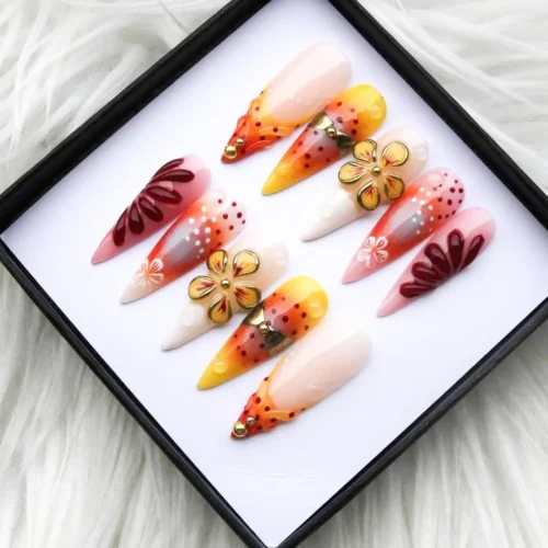 Orange and rose ombre press-on nails in a tilted black box on white fur, with gold flower charms and dotted tips