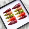 Angled view of tomato-themed press-on nails: dark red seed-slice nails, green plaid with vines and orange gradient nails
