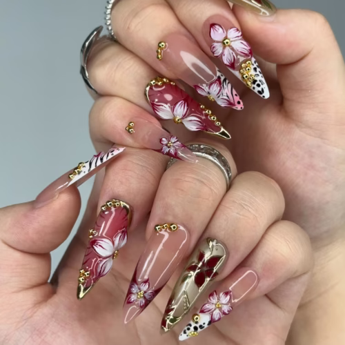 Burgundy and nude stiletto press-on nails worn on a hand: 3D flowers, gold beads, gold-foil tips and leopard spots