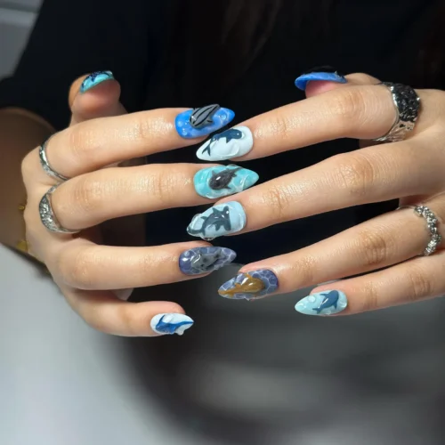 Blue, aqua and grey press-on nails worn on two hands with rings, each with a sculpted shark on a wave