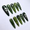 Long stiletto press-on nails in two greens with white skull-mask faces, black cobwebs and bat shapes, shown at an angle