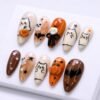 Almond press-on nails in cream, orange and brown on a white tray, with outlined ghost cats, a 3D pumpkin and a skull bow