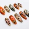 Almond press-on nails in orange, beige and black with cobwebs and spiders, bats, a gold skull charm and handwritten lettering
