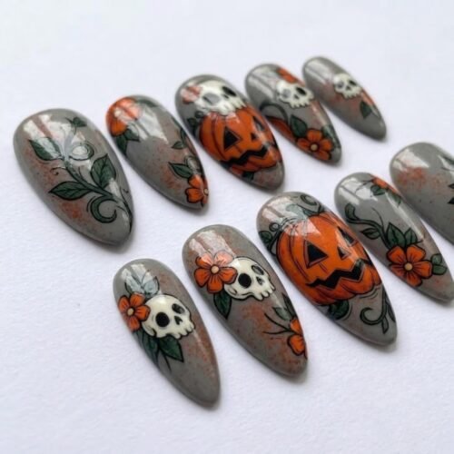 Angled view of gray almond press-on nails with big carved pumpkins, small skulls under orange flowers and green vines