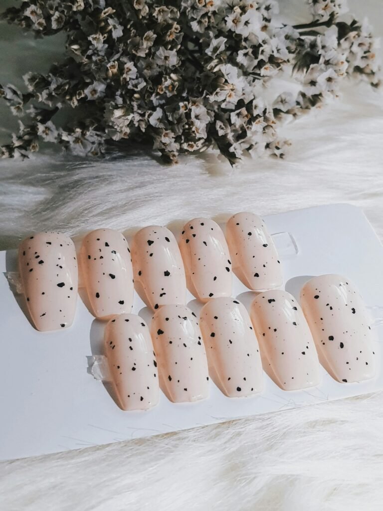 A handmade set of nude press-on nails with black speckles on a card next to dried flowers